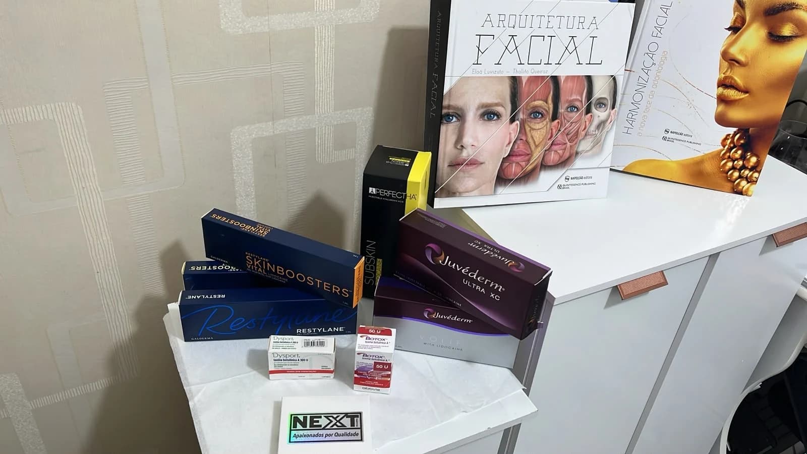 Embalagens de toxina botulínica Botox e Dysport, preenchedores Restylane, Juvéderm e Perfectha e Restylane Skinboosters sobre uma bancada, com livros de anatomia facial ao fundo